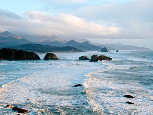 A scenic view of the ocean with waves crashing on rocky shores, surrounded by mountains under a cloudy sky.