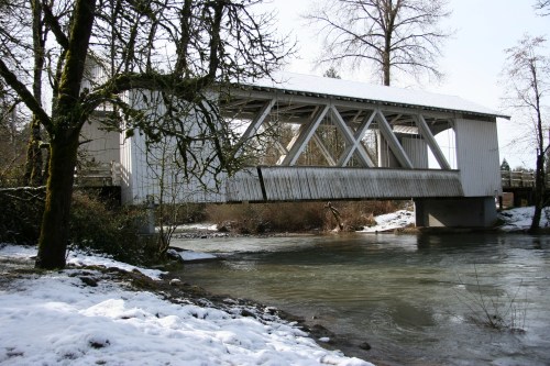 A white covered bridge spans a river, surrounded by snow-covered trees and a winter landscape.