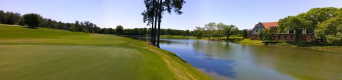 A panoramic view of a golf course with a lake, trees, and a building in the background under a clear blue sky.