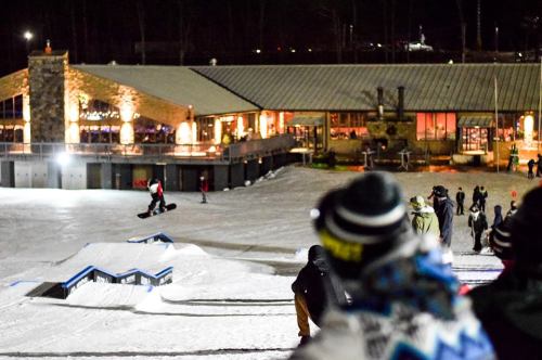 A snowboarder performs a trick at night, with a lit lodge and spectators in the background on a snowy slope.