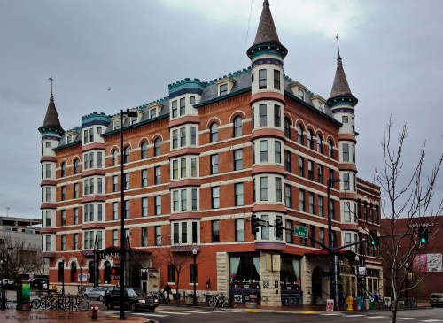Historic brick building with turrets, featuring colorful accents and a corner entrance, set against a cloudy sky.