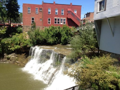 A serene waterfall cascades into a river, surrounded by greenery and buildings in the background.