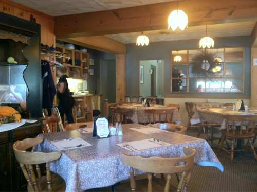 Cozy restaurant interior with wooden tables, chairs, and warm lighting; a staff member is seen in the background.