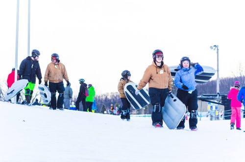 A group of people in winter gear walking up a snowy hill, carrying sleds and preparing for tubing.