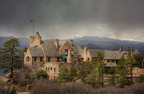 Historic stone mansion on a rocky hillside, surrounded by trees, with mountains and a cloudy sky in the background.
