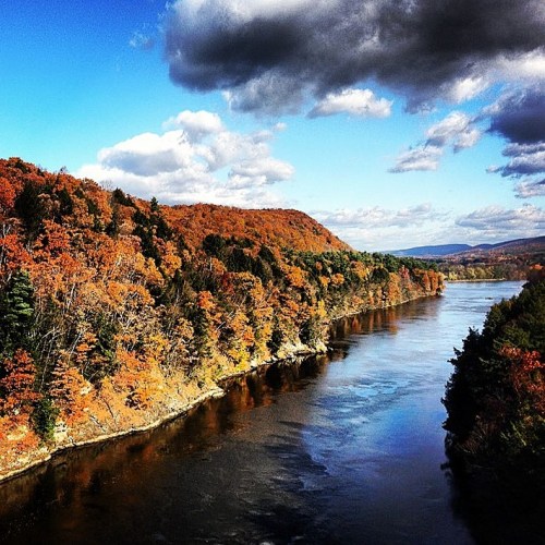 A serene river flows through a landscape of vibrant autumn foliage under a blue sky with scattered clouds.