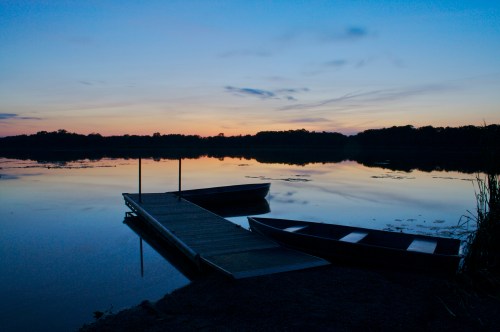 A serene lake at sunset, featuring a wooden dock and two boats, with calm water reflecting the colorful sky.
