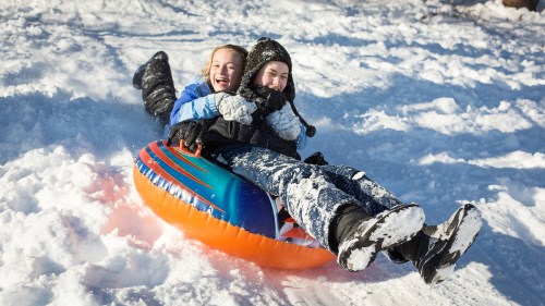 Two children joyfully sledding down a snowy hill on a colorful inflatable tube, surrounded by a winter landscape.