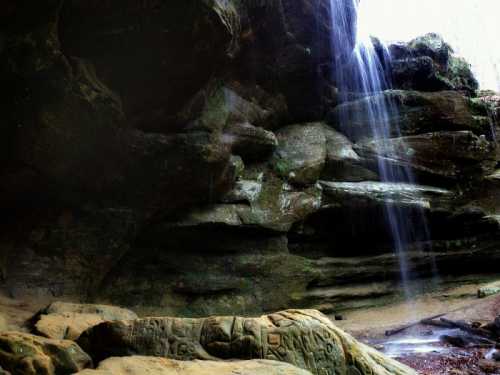 A serene waterfall cascades over rocky cliffs, surrounded by lush greenery and textured stone formations.