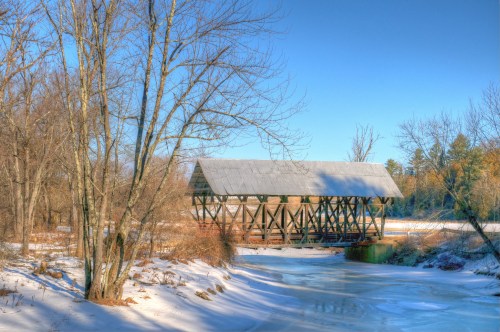 A rustic covered bridge stands over a frozen river, surrounded by bare trees and snow under a clear blue sky.