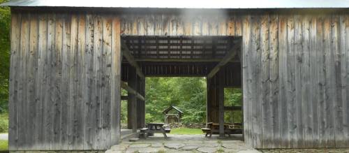 A rustic wooden structure with an open entrance, surrounded by greenery and picnic tables in a serene outdoor setting.