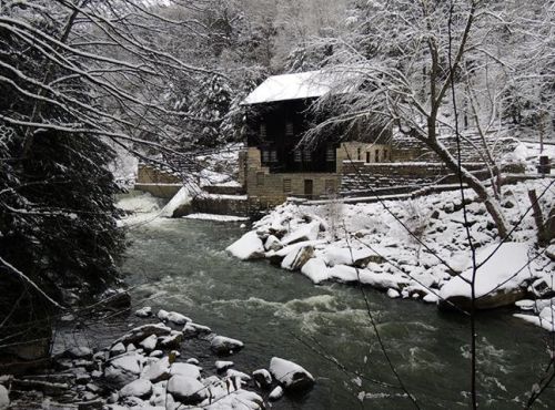 A snowy landscape featuring a river, rocks, and a historic building surrounded by trees.