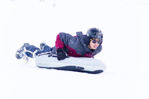 A person in a helmet and sunglasses sleds down a snowy hill, lying on a sled with a focused expression.