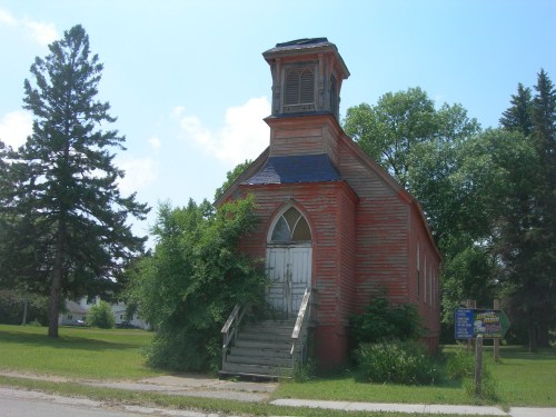An old, red wooden church with a steeple, surrounded by trees and overgrown grass, under a clear blue sky.