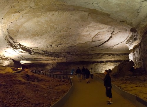 A spacious cave interior with a smooth ceiling, illuminated pathways, and visitors exploring the rocky terrain.