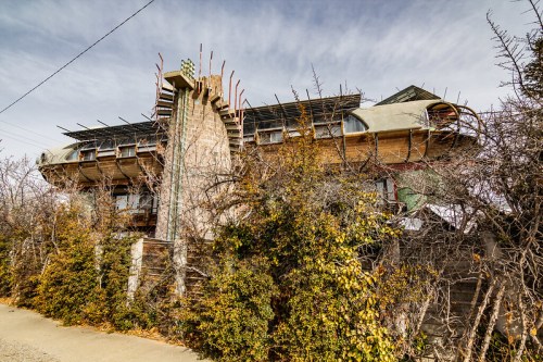 A unique, partially constructed building surrounded by overgrown vegetation and scaffolding under a cloudy sky.