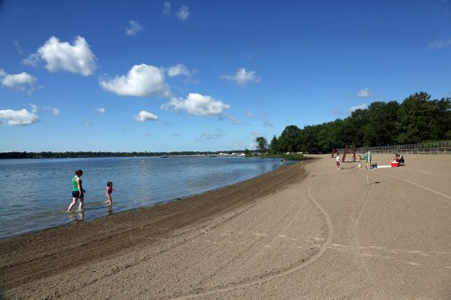 A sandy beach with people walking by the water, trees in the background, and a clear blue sky with clouds.