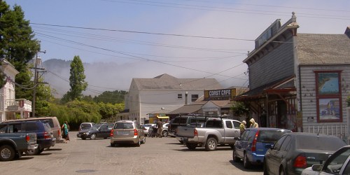 A small town street scene with parked cars, shops, and foggy mountains in the background.
