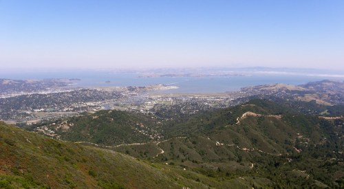 A panoramic view of a coastal city and bay, surrounded by green hills under a clear blue sky.