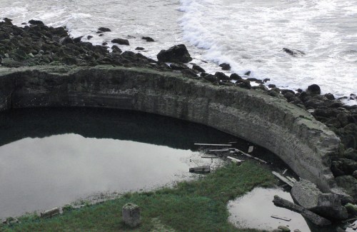 A curved stone structure partially submerged in water, surrounded by rocks and grass, with waves crashing nearby.