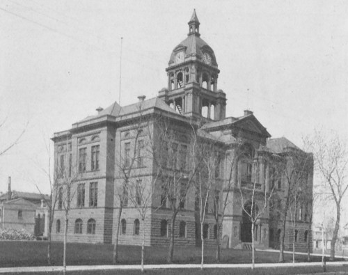 Historic building with a prominent clock tower, surrounded by bare trees and a clear sky. Black and white photo.