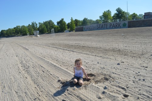 A child sits in a sandy area at a state park, surrounded by trees and a clear blue sky.