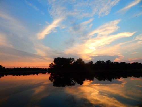A serene sunset over a calm lake, with colorful clouds reflecting on the water and silhouettes of trees in the foreground.