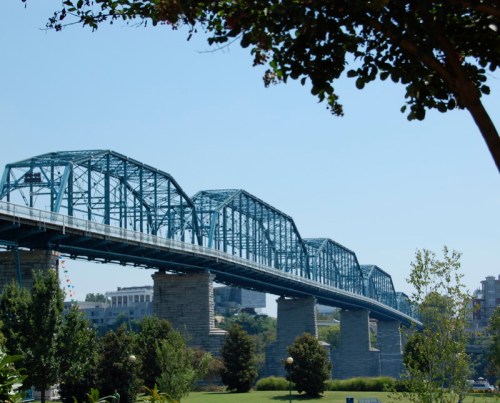 A blue steel bridge spans over a river, surrounded by greenery and a clear blue sky.