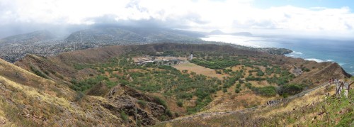 A panoramic view of a lush valley surrounded by mountains and ocean, under a partly cloudy sky.