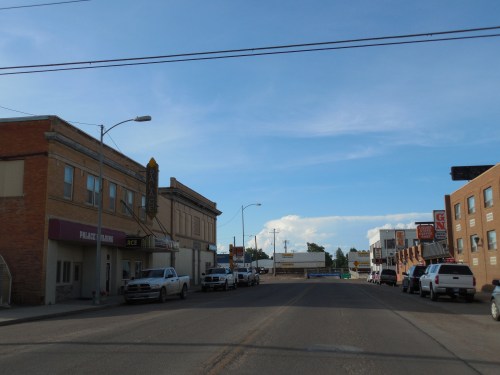 A small town street with brick buildings, parked cars, and a blue sky with scattered clouds.