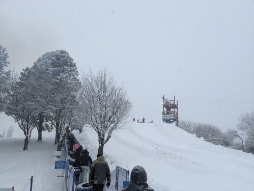 A snowy landscape with people lined up near a snow-covered hill and a wooden structure in the background.