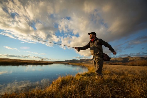 A person fishing by a river at sunset, casting a line with mountains and clouds in the background.