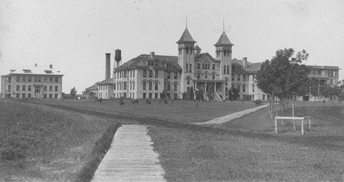 Historic black-and-white photo of a large, multi-story building with towers, surrounded by grass and a pathway.