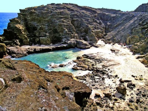 A rocky coastline with clear blue water and a sandy beach, surrounded by cliffs under a bright sky.