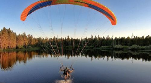 A colorful paramotor glides over a calm lake, surrounded by lush trees and a clear blue sky.