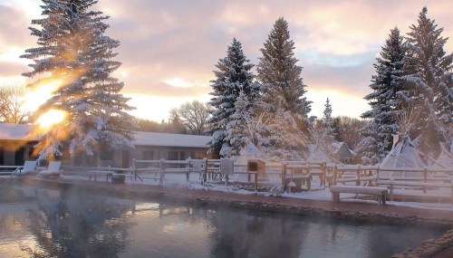 A serene winter scene with a steaming pool, snow-covered trees, and a sunrise in the background.