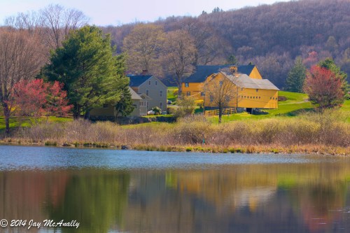 A serene lake reflects colorful houses and trees, surrounded by rolling hills and a clear blue sky.