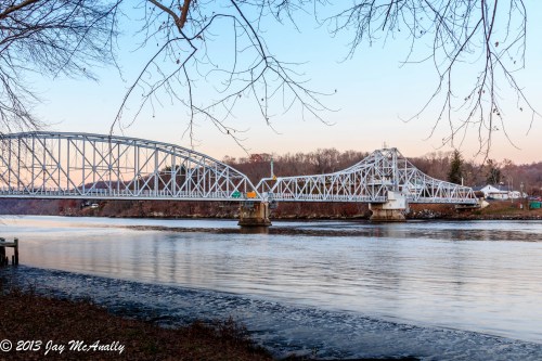 A white metal bridge spans a river, surrounded by trees and hills, under a soft blue sky at dusk.