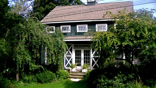 A charming green house surrounded by lush trees and plants, featuring a front porch and white trim.
