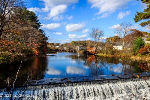 A serene river scene with a waterfall, surrounded by autumn foliage and reflecting clouds in the water.