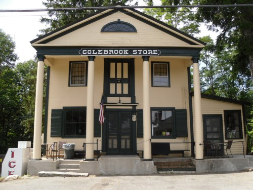 A two-story building with a sign reading "Colebrook Store," featuring columns and an American flag out front.