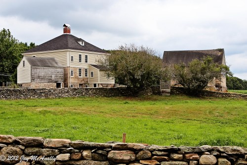 A large, two-story house with a stone wall in front, surrounded by green grass and trees under a cloudy sky.