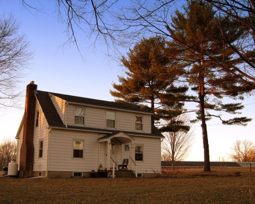 A two-story white house with a porch, surrounded by tall pine trees and a clear blue sky at dusk.