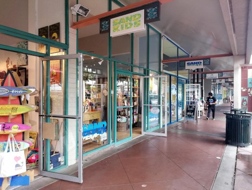 A colorful storefront with open doors, displaying surfboards and beach-themed merchandise in a shopping area.