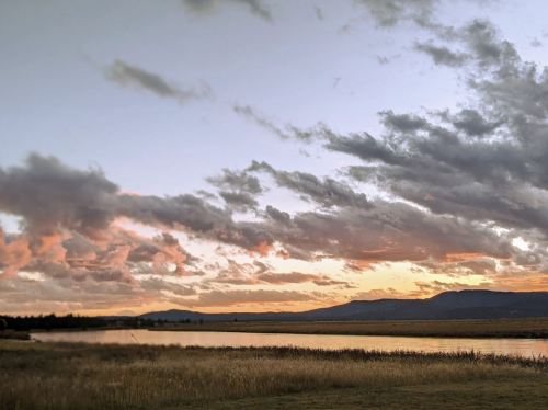 A serene landscape featuring a calm river, grassy banks, and a colorful sunset with clouds over distant mountains.