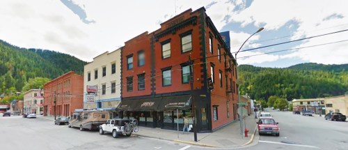 Historic brick building at a street corner, surrounded by mountains and parked vehicles.