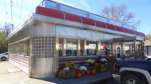 A retro diner with a red awning, large windows, and autumn decorations, including pumpkins and hay bales outside.