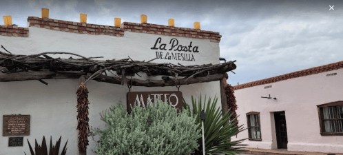 A rustic building with a sign reading "La Posta de la Mesilla," featuring adobe architecture and desert plants.
