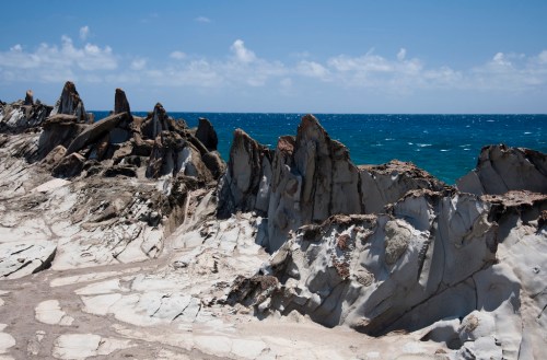 Rocky coastline with jagged formations under a clear blue sky and calm ocean waves in the background.