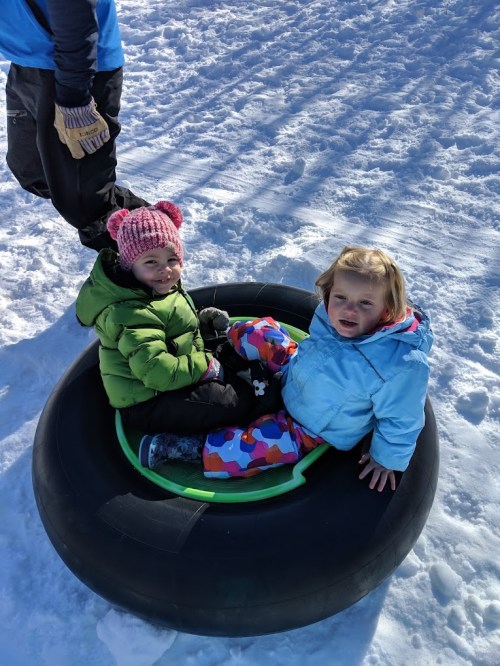 Two children sit in a snow tube, surrounded by snow, wearing colorful winter clothing and smiling.